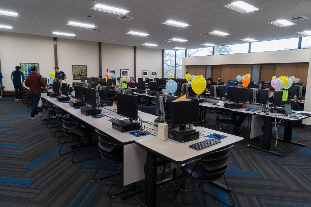 A computer lab with rows of desktop computers on long tables, each station decorated with colorful balloons. Several people are seated at the computers, working or talking, while a few individuals are standing or walking around.
